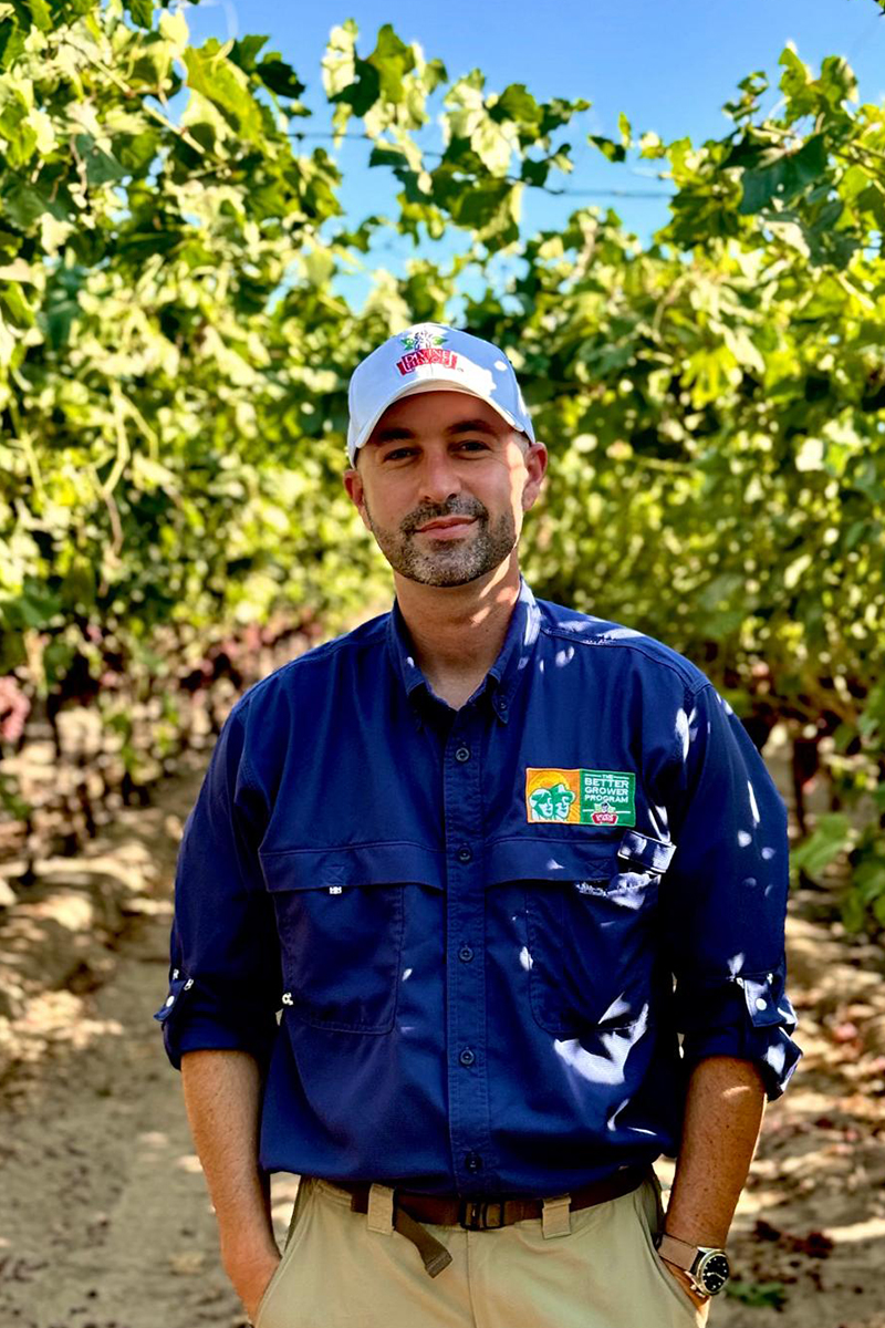 A man in a white baseball cap and a blue branded collared shirt stands in dappled shade in a vineyard