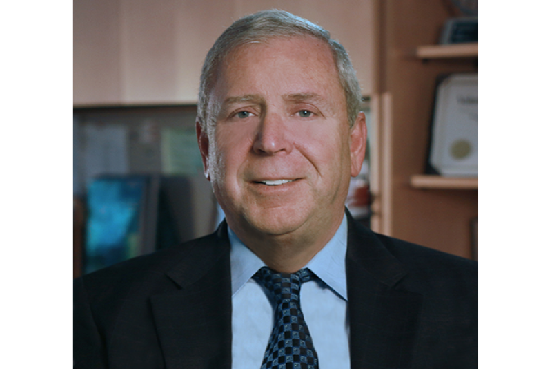 A portrait of an older middle-aged white man in a black suit, light blue shirt, and a green-checked tie.