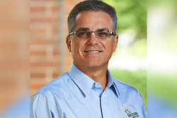 A portrait photo of a man with glasses who is wearing a light blue, button-up collared shirt. The background is out of focus, but shows a brick wall on the left of him and green vegetation to the right of him.