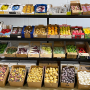A produce display of four racks with various items lining each one, plus boxes of mostly potatoes on the ground in front.