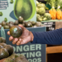 The arm of a shopper is in the photo, with an avocado in hand near a bin of avocados in a store