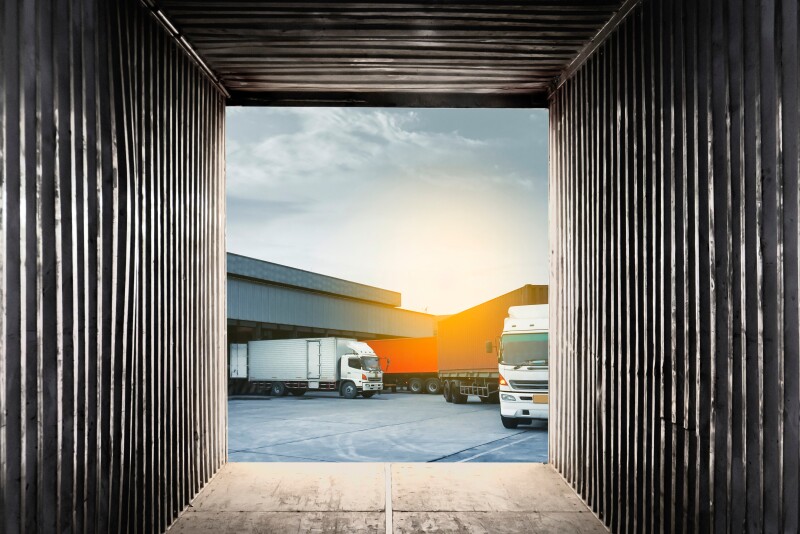 A view from inside a square, metal shipping container. Facing toward the outside, shown is part of a gray building with a semi truck backed into it on the left and another semi truck with a trailer on the right.