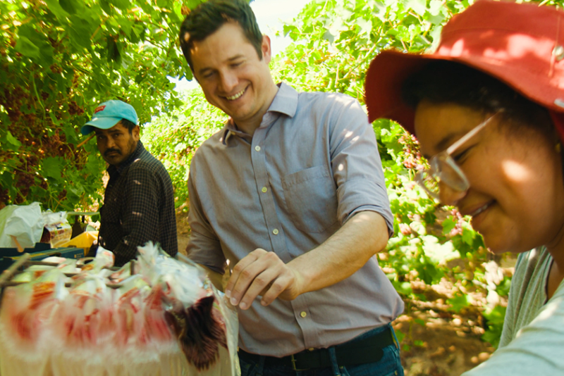 A man in the middle of the photo reaches for items in a basket. He is in a field, with another man to the left of the frame and a woman to the right.
