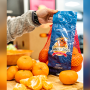 An arm coming from the left of the frame holds up a red-netted bag of clementines. The bag has a blue, vertical label with the Little Smoochies logo. In front of the bag are five loose clementines and one that has been peeled and split into pieces.