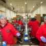 Two women in the foreground, each wearing a red jumpsuit and blue hair net, smile as they work on a packing line.