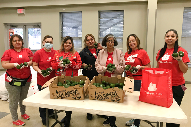 Seven women in red T-shirts stand side-by-side, holding avocados. There is a white table in front of them holding two cardboard boxes of avocados, next to a red tote with the Brighter Bites logo.