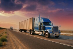 A large blue big rig hauling a full 53-foot dry van trailer along the highway, set against a colorful dusk skyline.