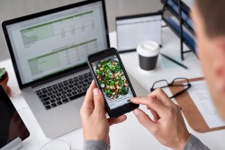 Man edits photo on his phone, sitting at a desk with his laptop in the background.