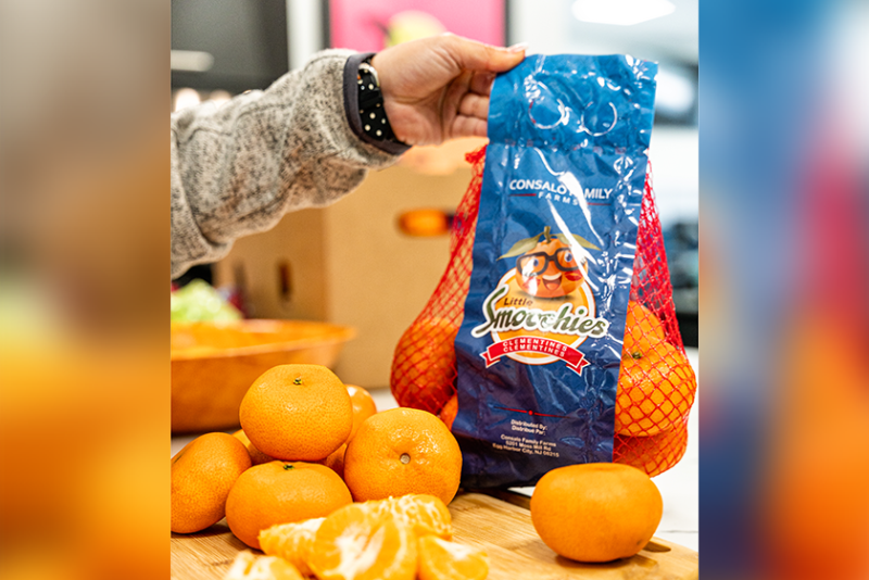An arm coming from the left of the frame holds up a red-netted bag of clementines. The bag has a blue, vertical label with the Little Smoochies logo. In front of the bag are five loose clementines and one that has been peeled and split into pieces.