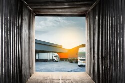 A view from inside a square, metal shipping container. Facing toward the outside, shown is part of a gray building with a semi truck backed into it on the left and another semi truck with a trailer on the right.