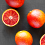 Six blood oranges on a black surface, seen from above. Two of the six are only halves, with the inside flesh facing the camera.