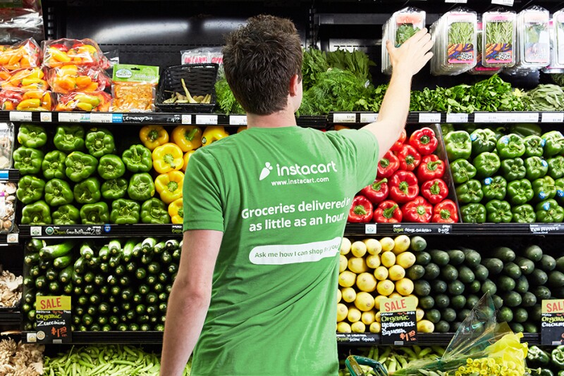 A person wearing a green Instacart shirt with their back to the camera stands in the produce department, reaching for some fresh herbs.