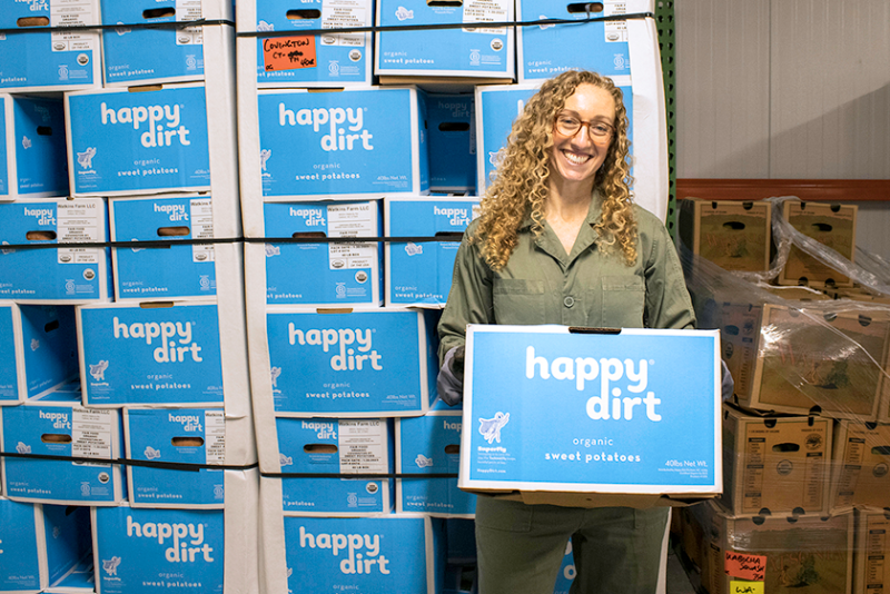 A woman is holding a box of sweet potatoes and standing in front of a wall of stacked boxes of sweet potatoes