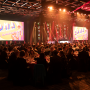A crowd of people sit at an array of round banquet tables in front of a stage with two large display screens