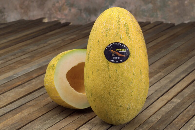A whole oblong yellow melon with a peacock sticker and a cut half of the same sort of melon, showing orange flesh, sit on a wooden-slatted table. 