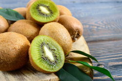 A close-up photo of a cluster of eight brown kiwifruit and two kiwifruit halves on a brown wooden cutting board, which is on a grayish-blue wooden tabletop.