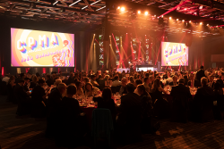A crowd of people sit at an array of round banquet tables in front of a stage with two large display screens
