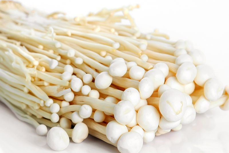 White enoki mushrooms, lying on their sides on a white surface and a white background. The mushrooms are stringy with small white caps.