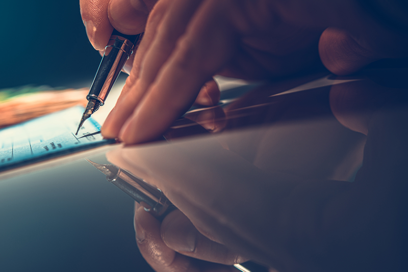 A hand is shown writing a check with a pen.