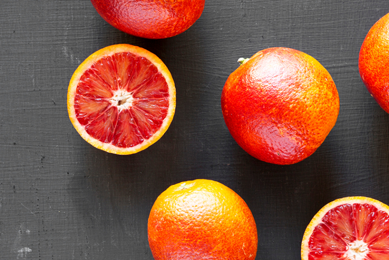 Six blood oranges on a black surface, seen from above. Two of the six are only halves, with the inside flesh facing the camera.