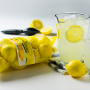 A yellow mesh bag of lemons next to a glass pitcher and cup of lemonade