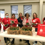 Seven women in red T-shirts stand side-by-side, holding avocados. There is a white table in front of them holding two cardboard boxes of avocados, next to a red tote with the Brighter Bites logo.