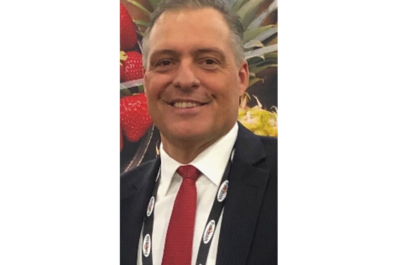 A smiling close up photo of a man in a suit with strawberries and a pineapple in the background.