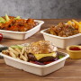 Shown are three containers: One in front holds french fries, pickles and a cheeseburger with lettuce, onion and tomato. The one on the back left holds sauced-covered Buffalo wings with celery and carrots. The one at middle right holds sauced boneless chicken fingers and french fries. There is a small yellow bowl holding sauce next to the containers, which are on a wooden tabletop.