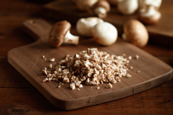 A professional photo of a rustic wooden cutting board on a wooden table. The focus is on a pile of diced mushrooms in the foreground, with whole white mushrooms out of focus in the background.
