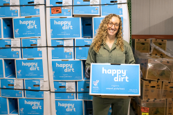 A woman is holding a box of sweet potatoes and standing in front of a wall of stacked boxes of sweet potatoes