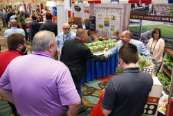 Two men shake hands at a booth on the trade show floor. They are surrounded by three other men and a woman, as well as other booths.