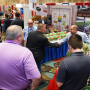 Two men shake hands at a booth on the trade show floor. They are surrounded by three other men and a woman, as well as other booths.