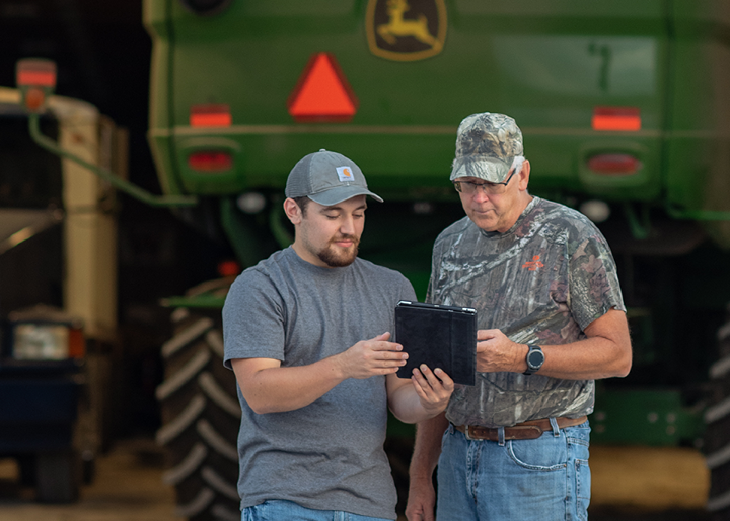 Two men with a tablet in front of a John Deere vehicle.