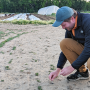 A man wearing a blue ball cap, black jacket and tan pants squats down to pick up bird feces from the ground.