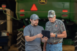 Two men with a tablet in front of a John Deere vehicle.