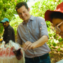A man in the middle of the photo reaches for items in a basket. He is in a field, with another man to the left of the frame and a woman to the right.