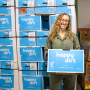 A woman is holding a box of sweet potatoes and standing in front of a wall of stacked boxes of sweet potatoes