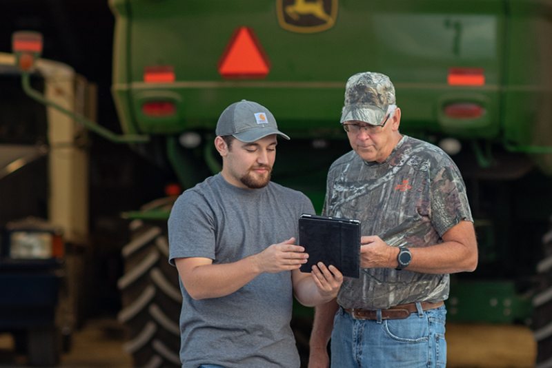 Two men with a tablet in front of a John Deere vehicle.