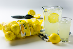 A yellow mesh bag of lemons next to a glass pitcher and cup of lemonade