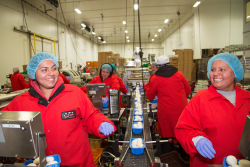 Two women in the foreground, each wearing a red jumpsuit and blue hair net, smile as they work on a packing line.