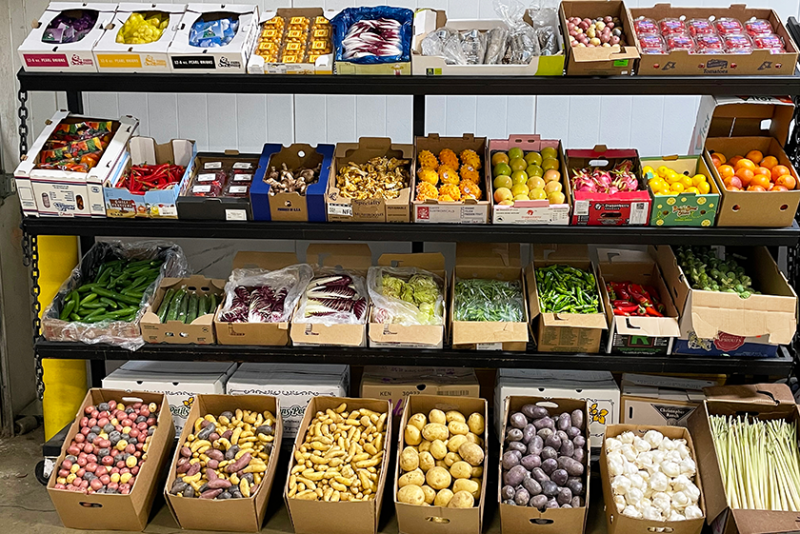 A produce display of four racks with various items lining each one, plus boxes of mostly potatoes on the ground in front.