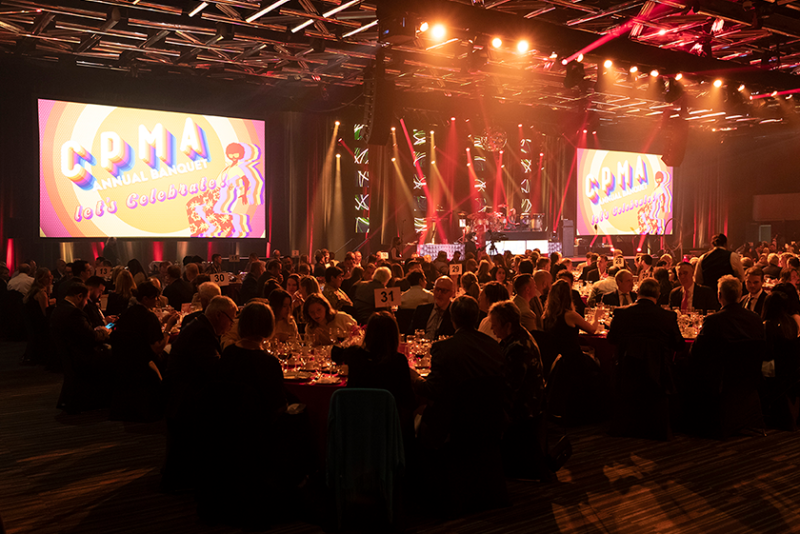 A crowd of people sit at an array of round banquet tables in front of a stage with two large display screens