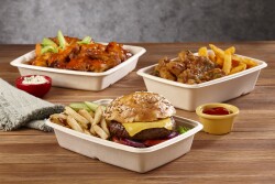 Shown are three containers: One in front holds french fries, pickles and a cheeseburger with lettuce, onion and tomato. The one on the back left holds sauced-covered Buffalo wings with celery and carrots. The one at middle right holds sauced boneless chicken fingers and french fries. There is a small yellow bowl holding sauce next to the containers, which are on a wooden tabletop.