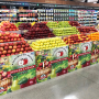 A view of a produce department display of several stands holding apples of different varieties, red and green. The four stands in front are identical, each bearing the Sage Fruit Co. logo and a photo of a basket of apples and a pear.